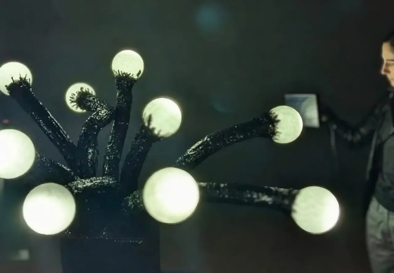 A person stands beside a dark branching sculpture with glowing spheres, holding a small device toward the lights